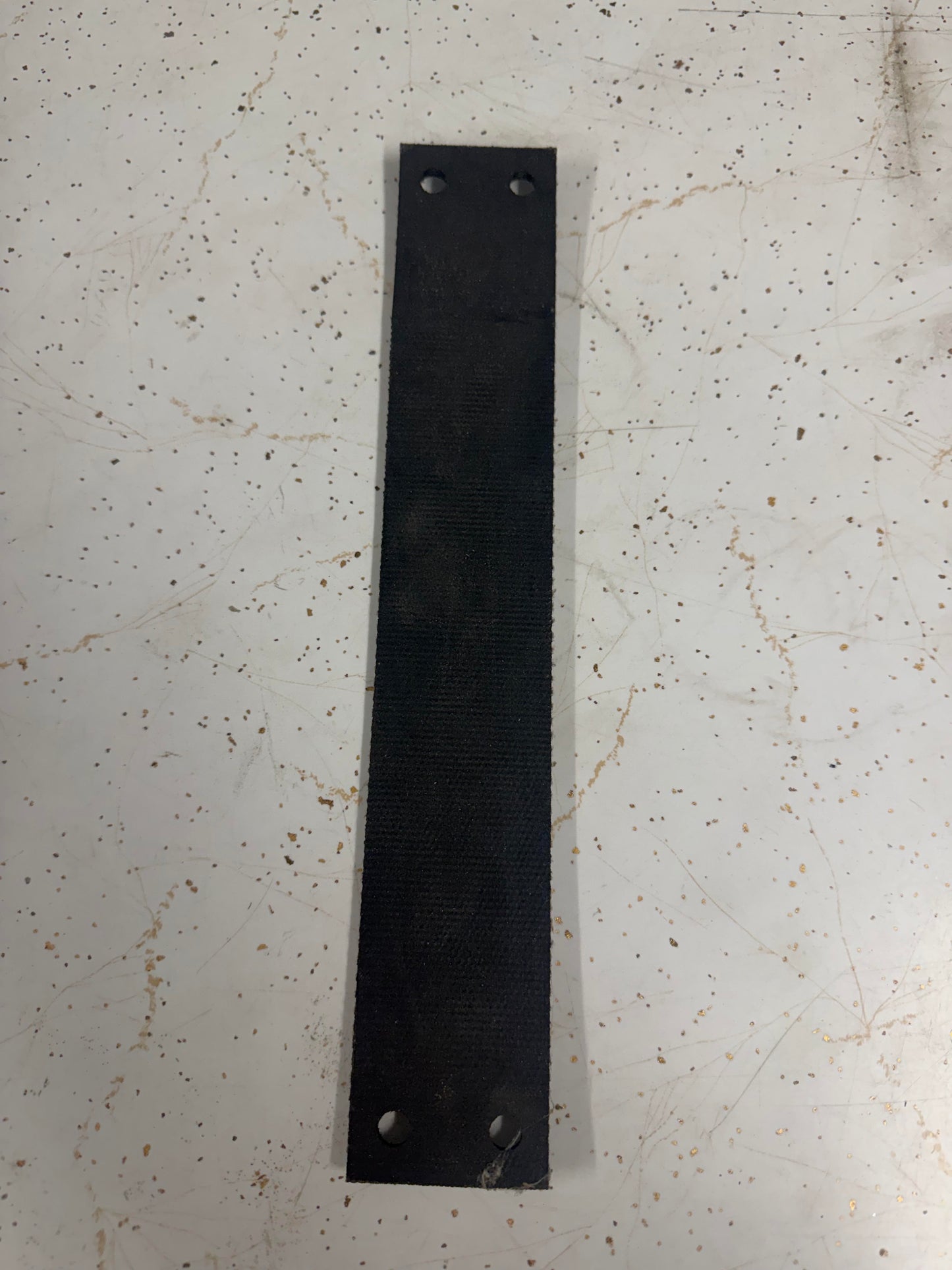 Photograph of a long black textured industrial rubber strap featuring four circular mounting holes and a fiber-reinforced surface lying on a white speckled and scratched countertop